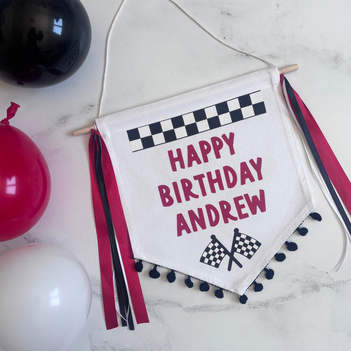 
                  
                    personlalized racing theme Happy Birthday banner with checkered flags and pom-poms on a marble surface with balloons.
                  
                
