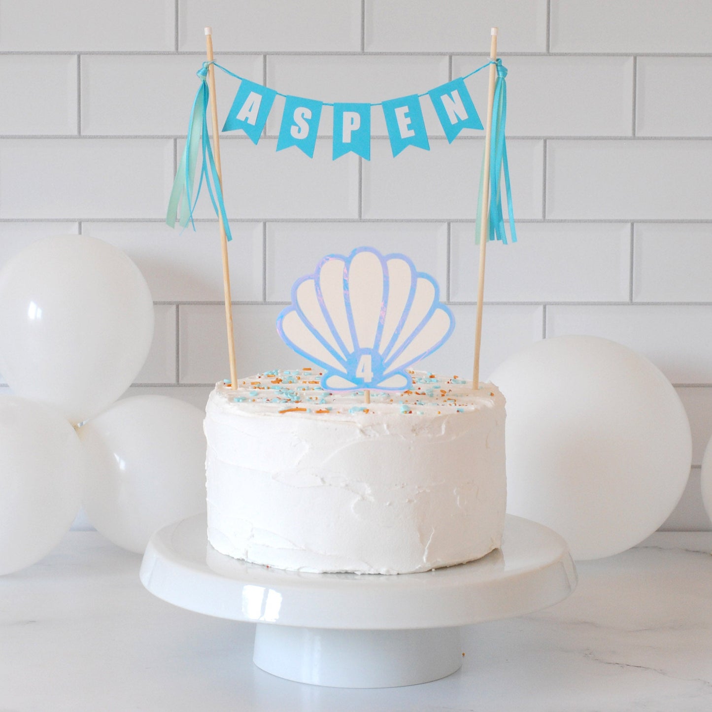 
                  
                    A white frosted birthday cake topped with a seashell-shaped topper and a blue banner reading ASPEN, set against a white subway tile wall with white balloons.
                  
                