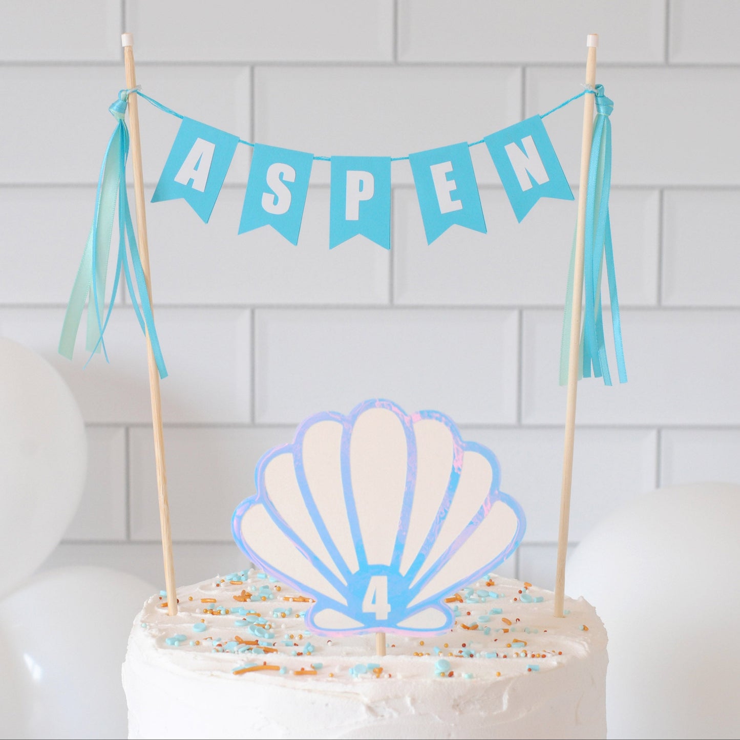 
                  
                    A white frosted birthday cake topped with a seashell-shaped topper and a blue banner reading ASPEN, set against a white subway tile wall with white balloons.
                  
                