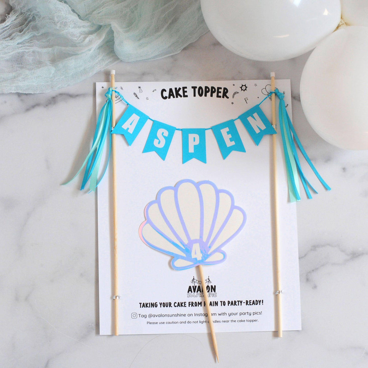 
                  
                    Under the sea cake topper set with cake banner with the name 'ASPEN' on blue flags, wooden sticks, turquoise tassels, and a seashell graphic on a white card.
                  
                