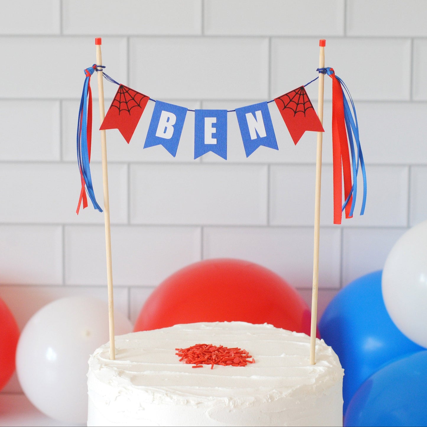 
                  
                    spiderman theme cake topper personalized with name banner in blue with white letters and red flags on each end with spider webs. Shown on a white cake with red sprinkles and balloons in the background
                  
                