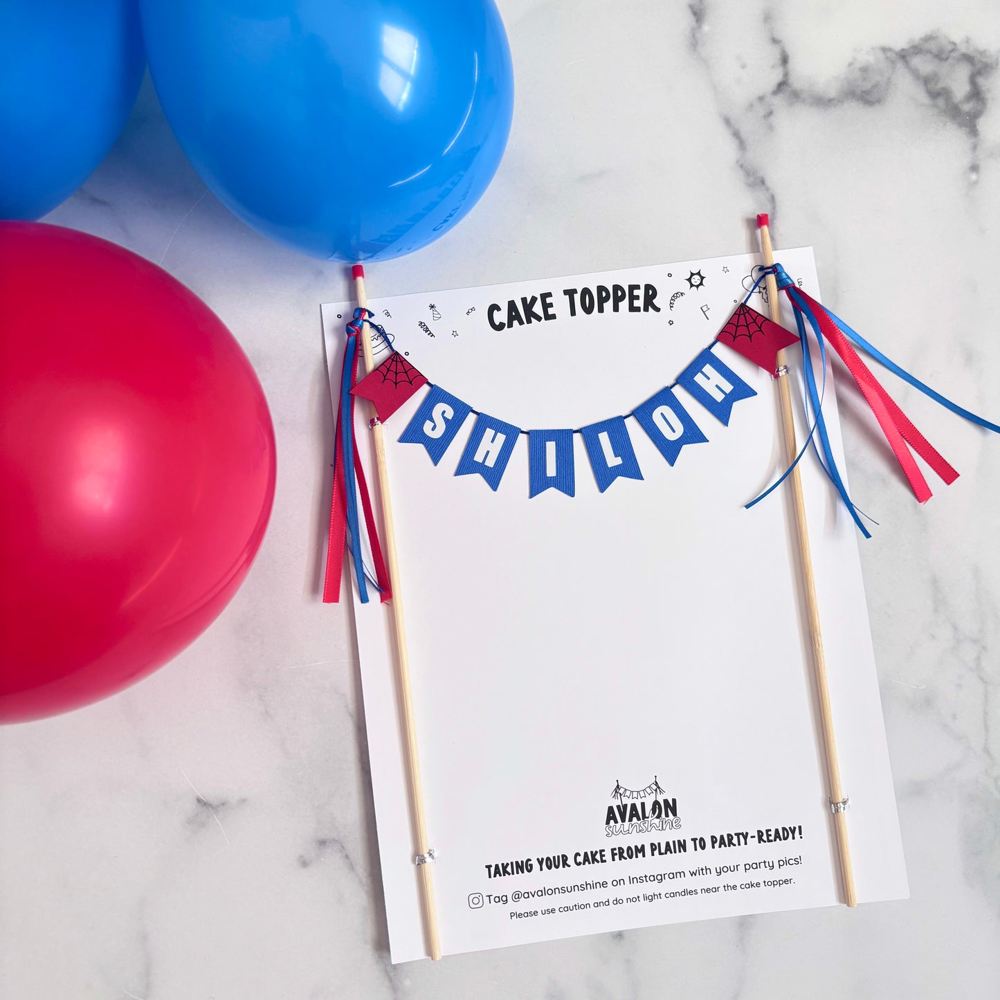 
                  
                    Spiderman cake topper personalized with name 'SHILOH'.  Blue name banner with white letters and red and black spiderweb flags on each end.  Shown in packaging on a marble countertop
                  
                