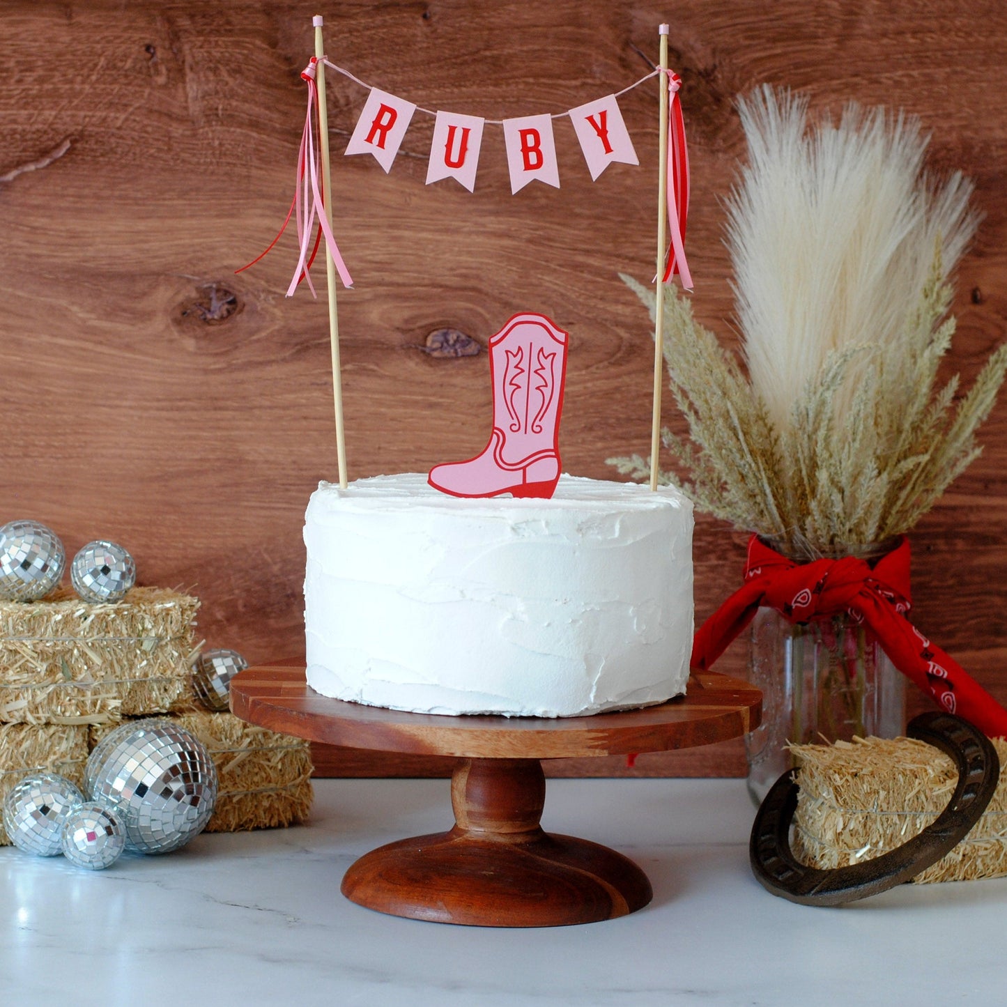 
                  
                    A white frosted cake on a wooden pedestal stand, decorated with a pink cowboy boot topper and a string banner reading RUBY. The scene is styled with hay bales, disco balls, a vase of dried grass with a red bandana, and a horseshoe against a rustic wood plank background.
                  
                