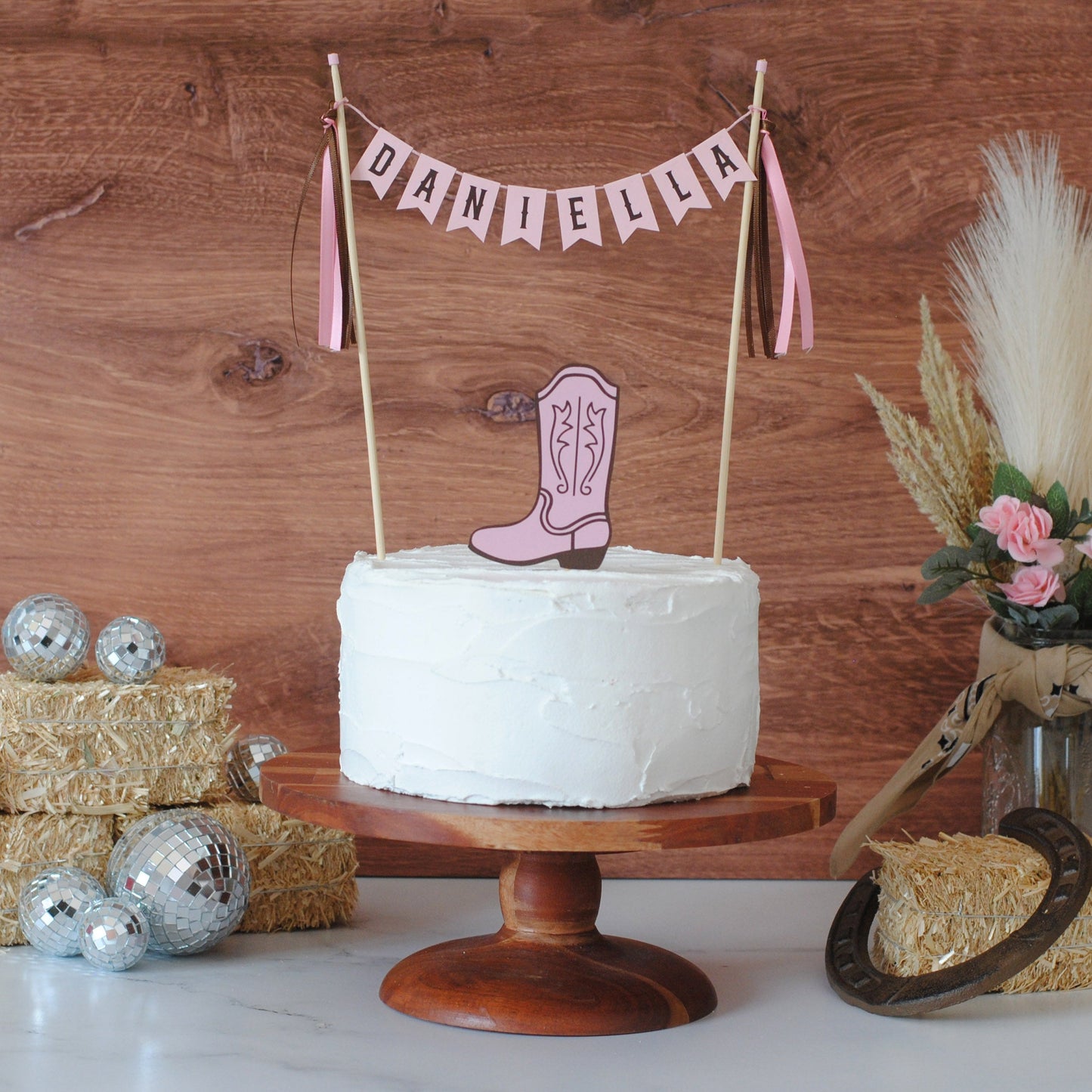 
                  
                    A white-frosted round cake on a wooden pedestal stand, decorated with a pink cowboy boot topper and a pink banner reading 'Daniella' against a rustic wood plank background. Hay bales, disco balls, and a small floral arrangement with dried grasses are styled around the cake.
                  
                