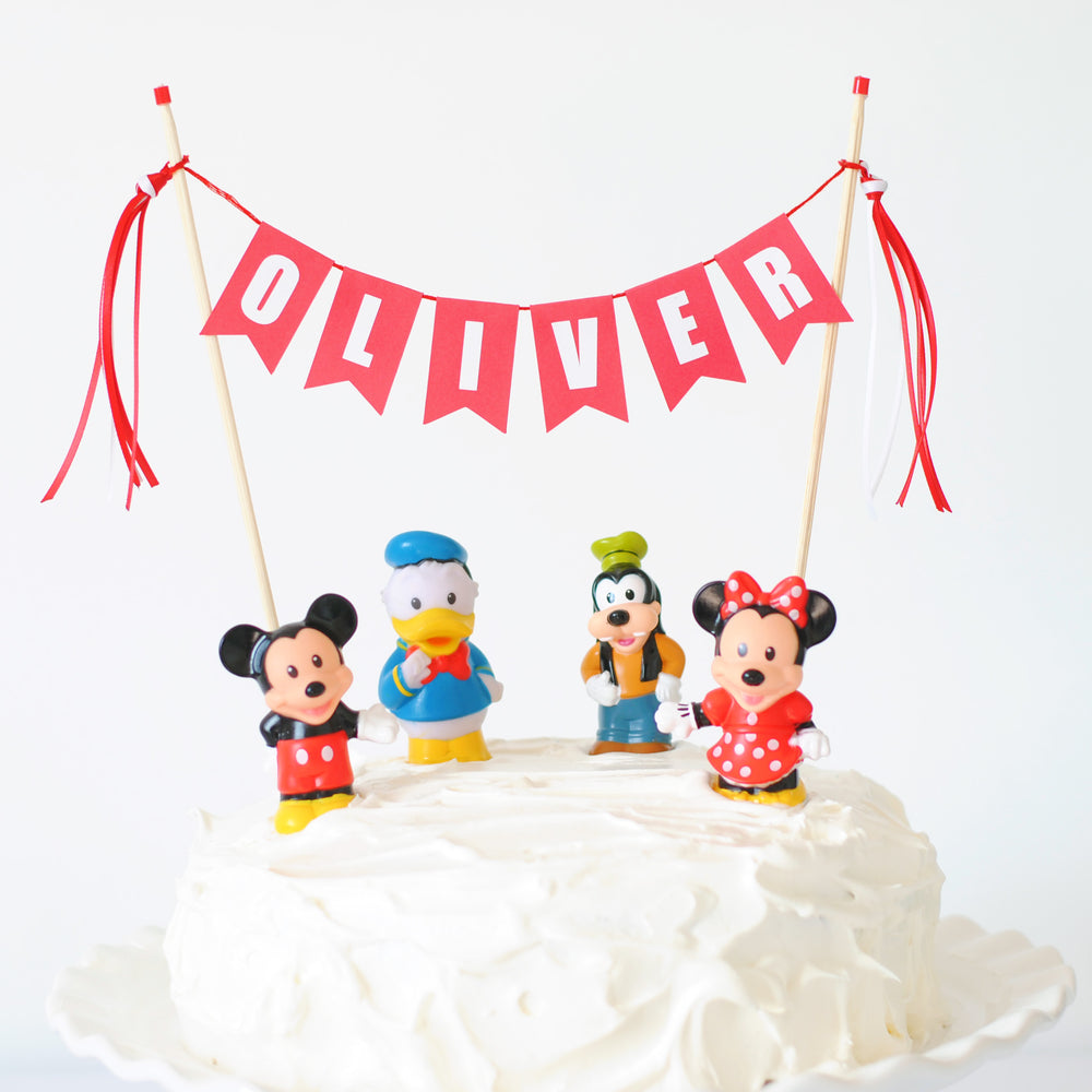 
                  
                    red and white name cake topper on a white cake shown with mickey and minnie mouse toys on the cake
                  
                