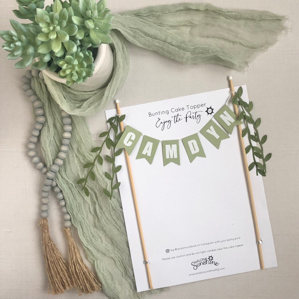 
                  
                    Personalized name cake topper in sage green with white letters and green leafy ribbons on the ends
                  
                