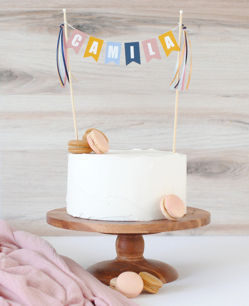 
                  
                    plain cake with french macarons and a muted rainbow color name cake topper
                  
                