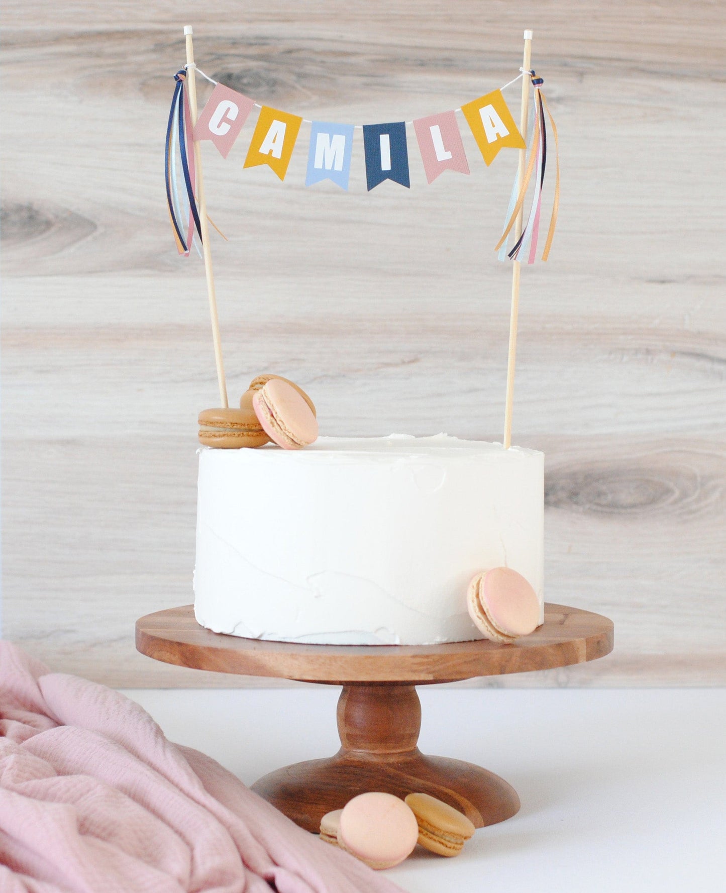 
                  
                    plain cake with french macarons and a muted rainbow color name cake topper
                  
                