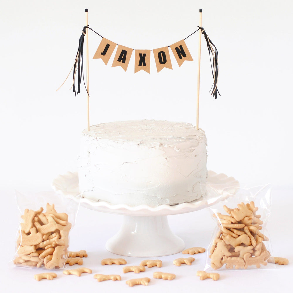 
                  
                    Kraft neutral cake banner with black letters personalized with name.  Shown with cake and animal crackers. 
                  
                