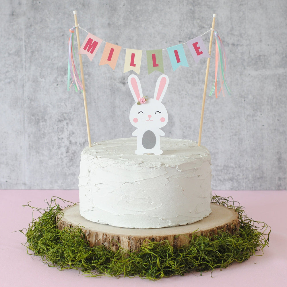 pastel rainbow name banner personalized with name.  Paper bunny cake topper underneath with a flower on its ear.