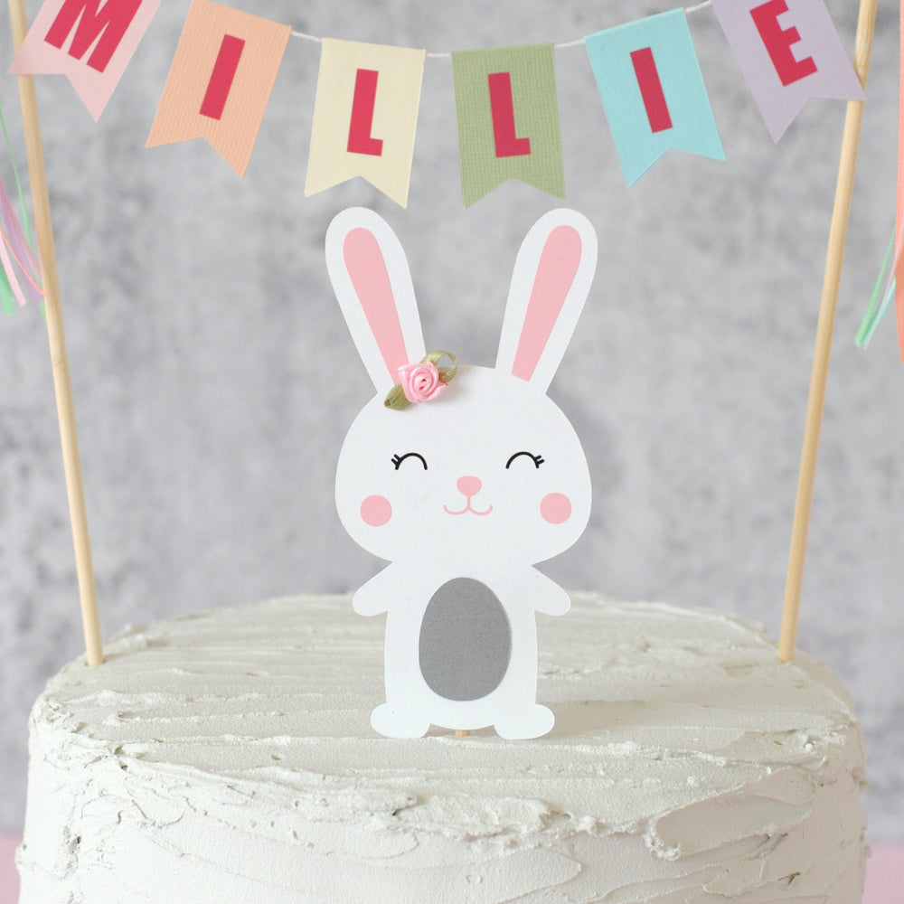
                  
                    pastel rainbow name banner personalized with name.  Paper bunny cake topper underneath with a flower on its ear.
                  
                