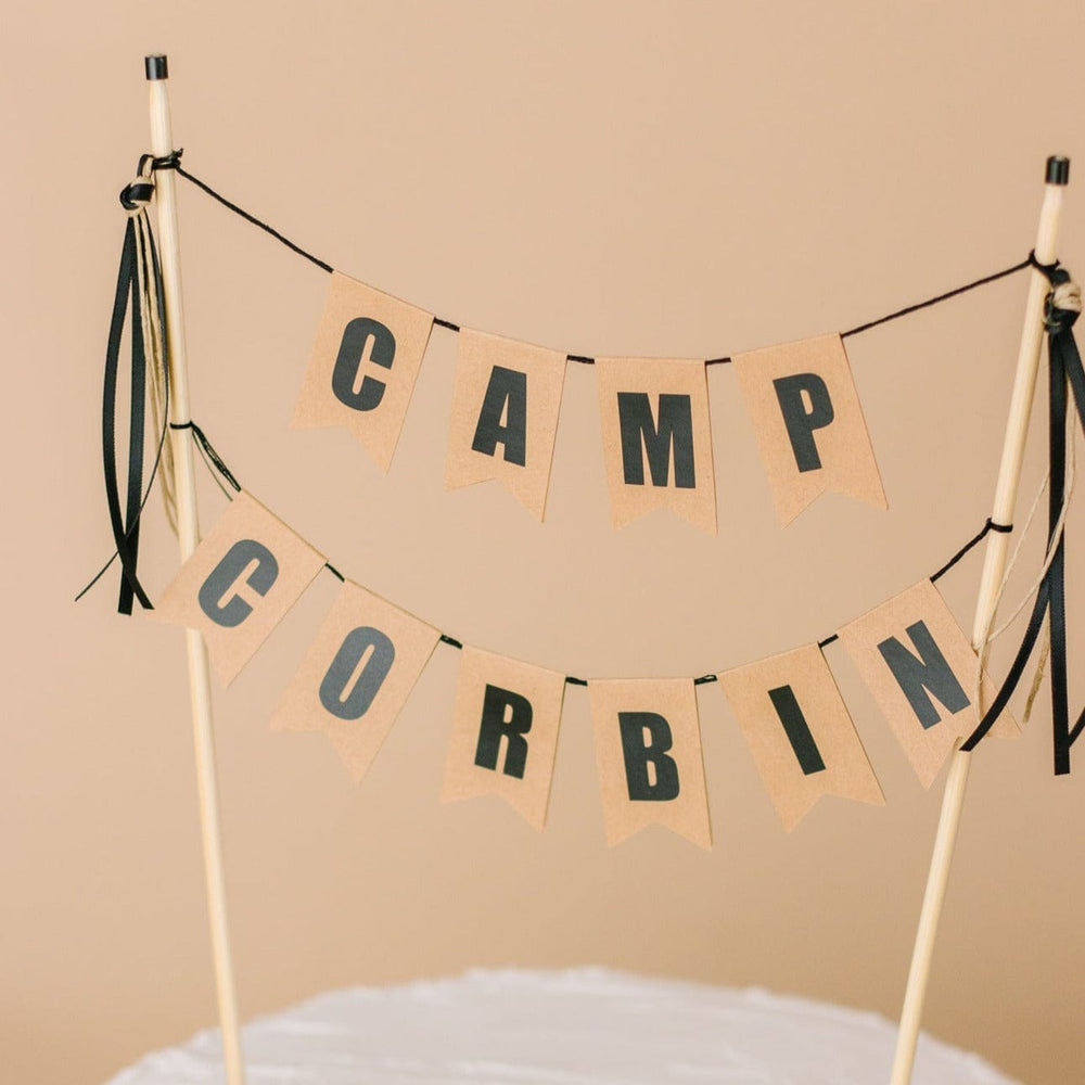 
                  
                    Two tier cake topper for camping theme birthday party.  Brown paper flags and black letters. top row says "camp".  Bottom row is personalized with a name.  
                  
                