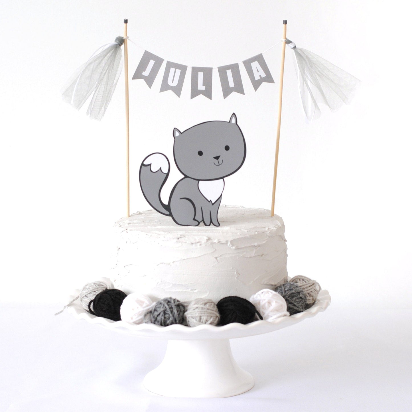 
                  
                    grey cat cake topper with matching name banner on top of the cake.  Cake is accessorized with mini yarn ball decorations
                  
                