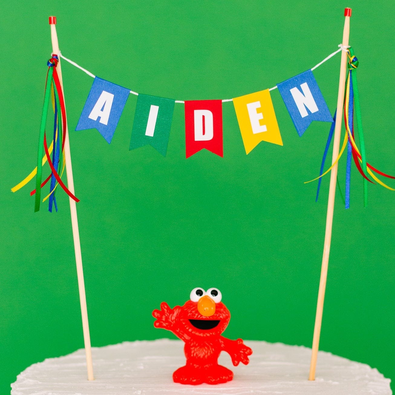 multi-color personalized name cake topper in blue, green, red and yellow shown with an Elmo figure on top of the cake