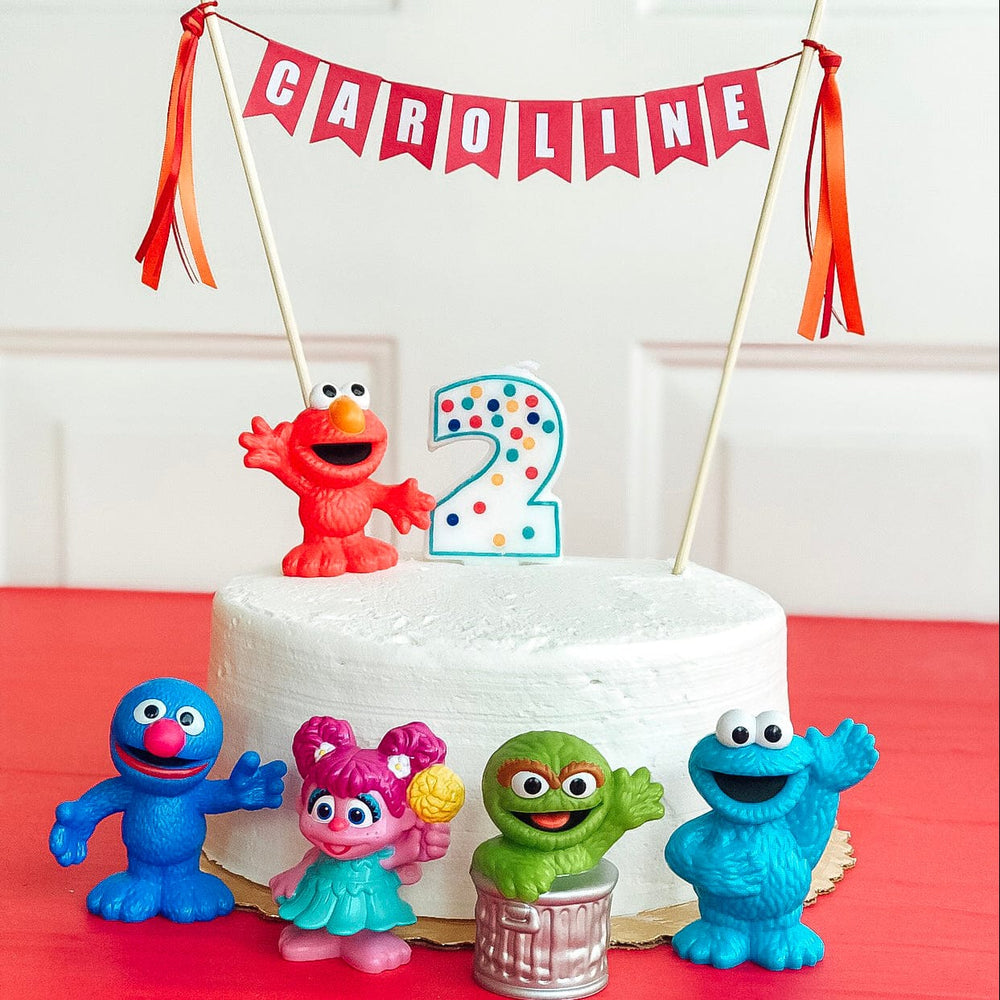 
                  
                    personalized birthday cake topper for kids with name in red and white, shown with sesame street toys
                  
                