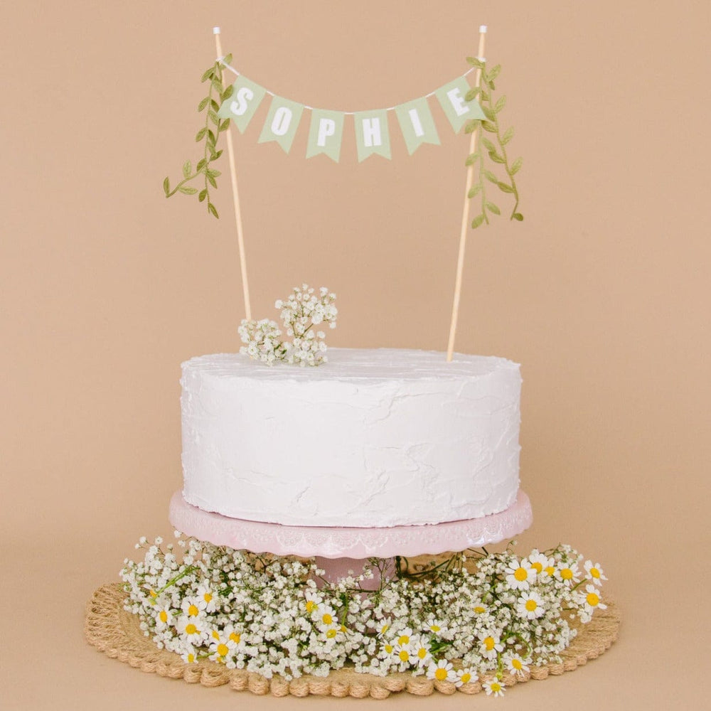 Greenery themed personalized cake topper with name and leaf ribbons, shown on a white cake decorated with small flowers