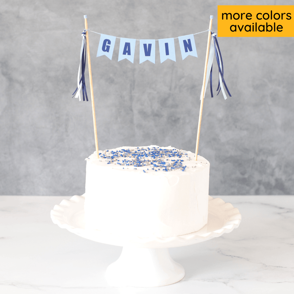 light blue cake banner with dark blue letters personalized with name and matching ribbon tassels shown on a white cake  with sprinkles
