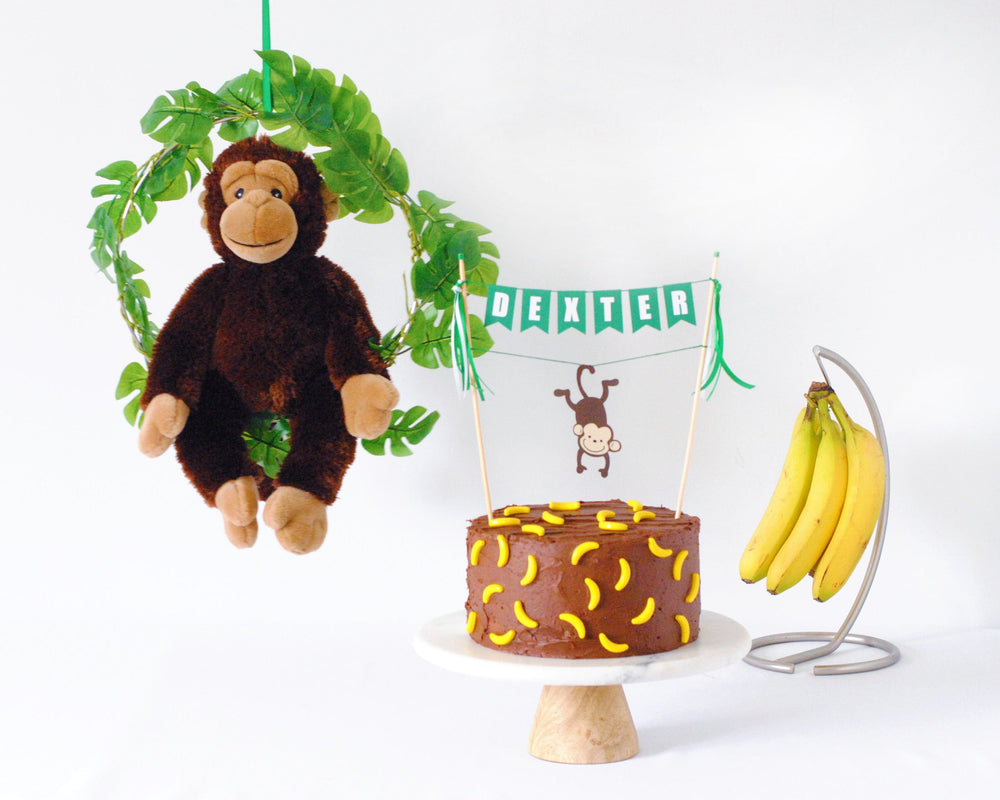 
                  
                    monkey cake topper on a chocolate cake with banana candies, bunch of bananas in the background and a hanging monkey decoration
                  
                