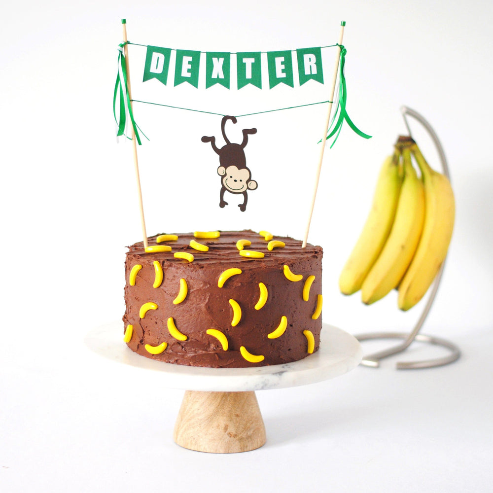 monkey cake topper with name banner and a monkey hanging from its tail over a chocolate cake with banana candy decorations