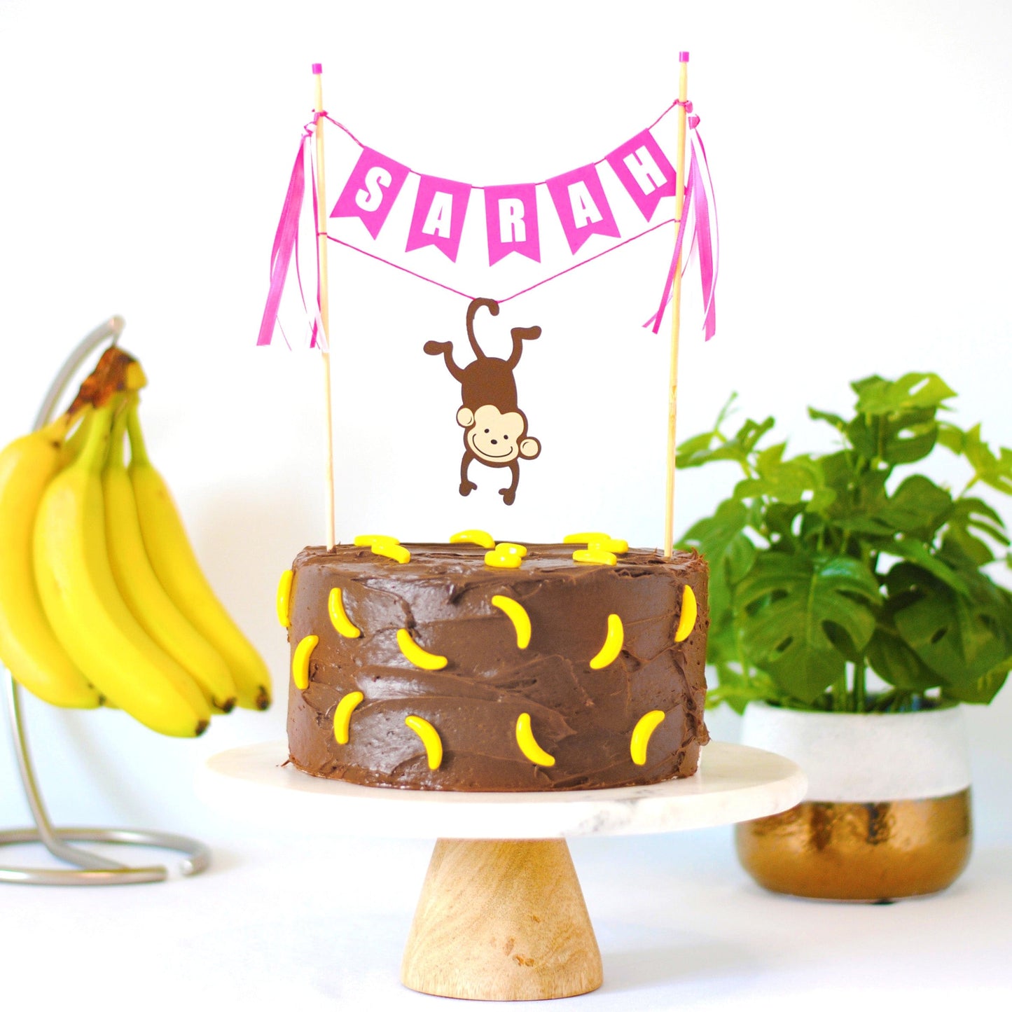 girls monkey birthday cake topper with name banner and monkey hanging from its tail shown on chocolate frosted cake with banana candies