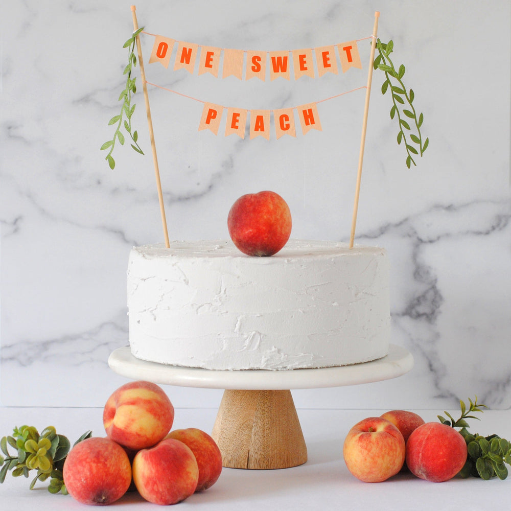 ONE SWEET PEACH two tier cake banner with green leafy tassels shown on a white cake with peaches on the table