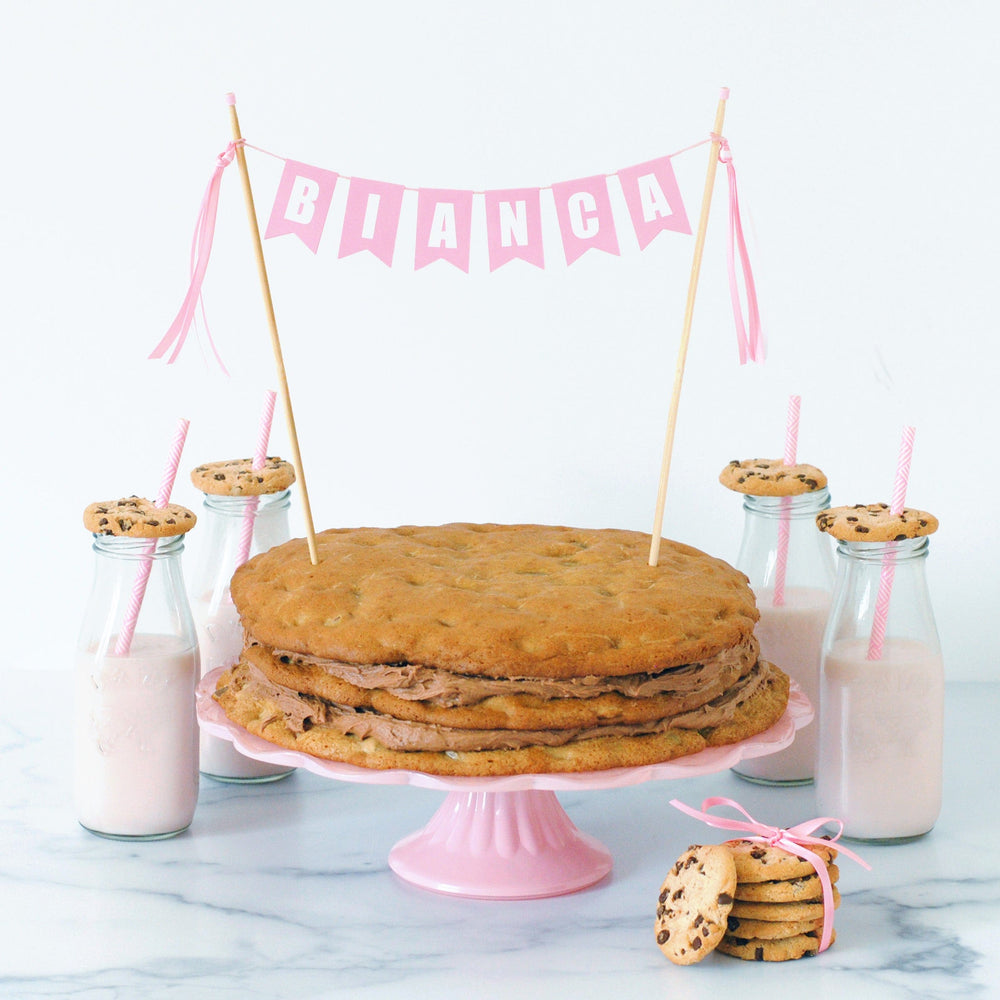 
                  
                    pastel pink birthday cake topper for girls, name banner shown on a layered chocolate chip cookie cake
                  
                