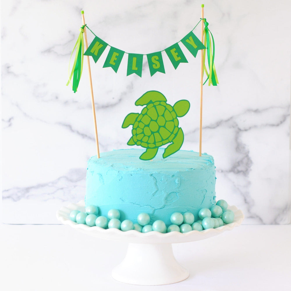 Sea Turtle themed cake topper set with personalized name banner in shades of green and a paper sea turtle on a cake